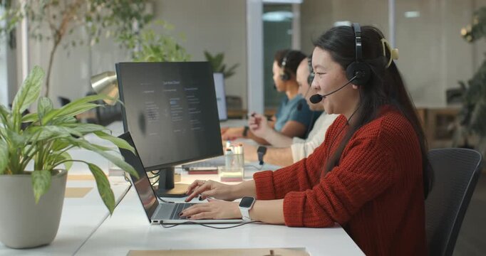 Diverse coworkers with Asian woman in red sweater taking call, typing on laptop at shared desk