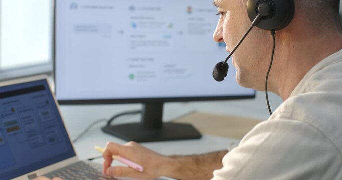 Receiving call, man at desk typing on laptop, pointing pink pen, speaking into headset for support