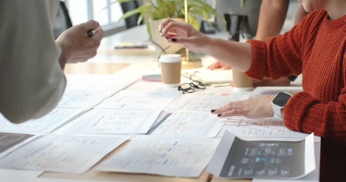 Team at office pen-cue urging Asian woman orange knit sliding, pointing mockups on table to review