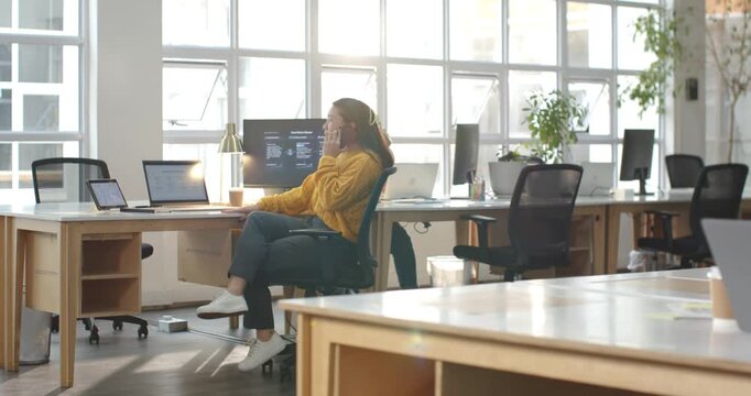 Woman in yellow sweater receiving call, holding phone, checking laptop and screen at office working
