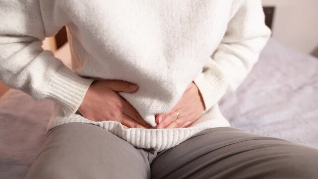 Young woman holding lower abdomen while sitting on bed. Slow motion footage illustrates painful intestinal cramps and digestive discomfort.