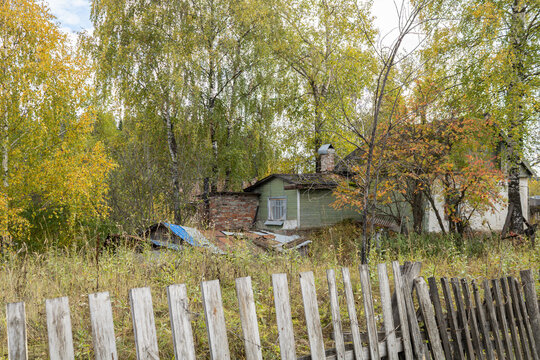 An old abandoned residential private house behind a crooked fence and colorful autumn trees in a provincial town of Russia.