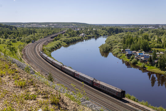 Long railway with RZD train traveling beside a winding river and private residential houses in summer.