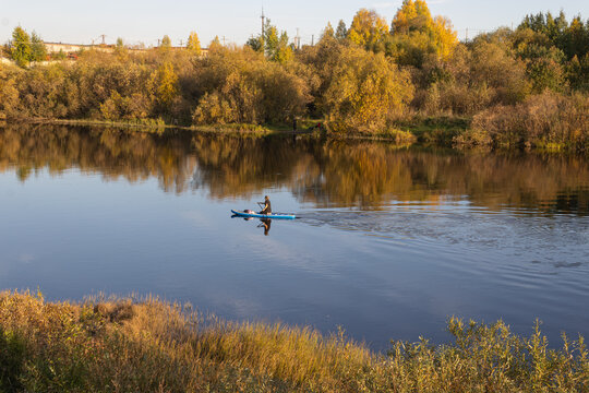 A woman paddling sitting on SUP along a calm river reflecting autumn forest in a provincial town of Russia.