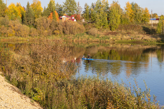 Serene river and autumn forest with woman paddling on SUP by private houses on shore in provincial town of Russia.