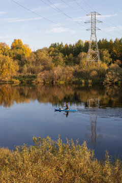 Serene river with a woman paddling on SUP board by an autumn forest in a provincial town of Russia.