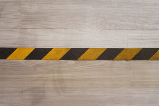 Close-up top view of a black and yellow diagonal striped safety caution tape marking a line on a light wood-grain laminate floor, indicating a restricted area, social distancing boundary, or hazard