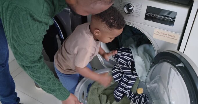 African American father guiding child, child pulling clothes from basket into dryer, starting cycle