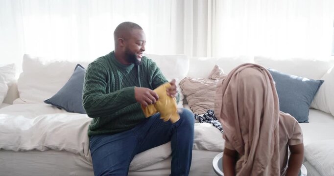 African American father and toddler son folding shirts on sofa as child reaching into laundry bin