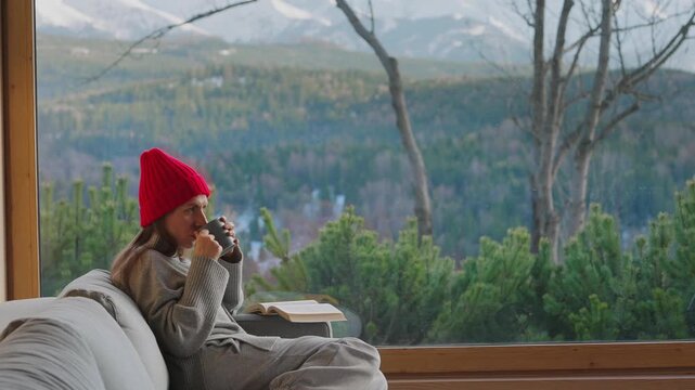 A woman reading by a large window and enjoying scenic snowy mountain view.