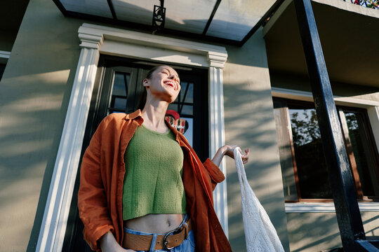 Happy young woman with shopping bag laughing near house entrance. Low angle view of a joyful girl in green top and orange jacket. Concept of lifestyle, happiness, and leisure.