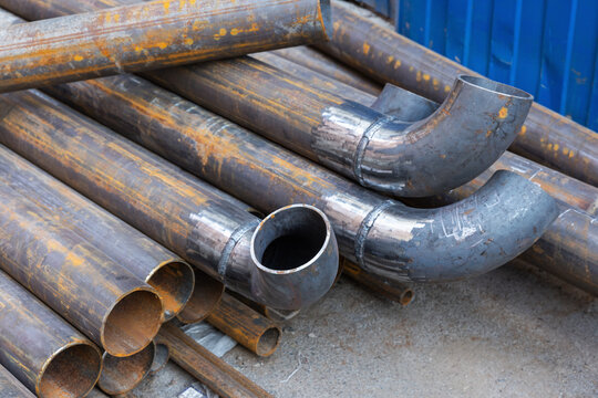 Stacked steel pipes and welded elbow fittings showing rust, weld marks and industrial texture at a construction yard. High-detail view of metal tubing, fabrication work, and raw manufacturing material