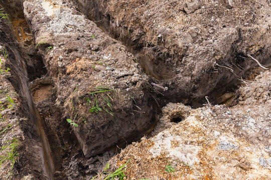 Deep excavation trenches cut into clay and sandy soil at a construction site, showing exposed roots, uneven earth, and drainage channels ready for foundation, utility work, or landscaping preparations