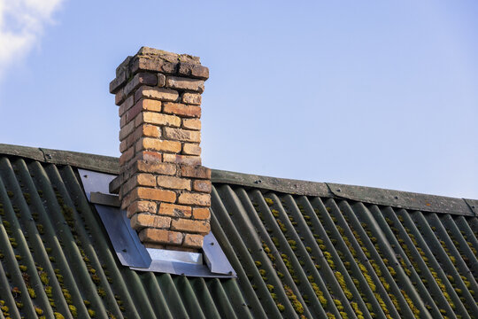 A weathered brick chimney rises from a green corrugated roof dotted with moss, set against a clear blue sky. Rustic rooftop detail highlighting texture, age, and outdoor architectural character