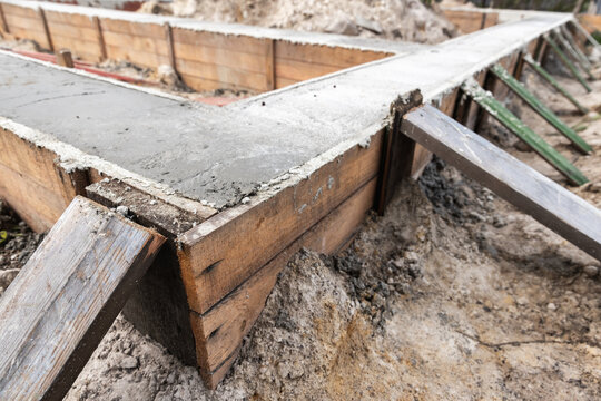 Concrete foundation pour inside wooden formwork with angled braces at a residential construction site, showing fresh cement, dirt and construction materials during early building and groundwork phase
