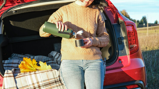 Woman pouring hot tea from thermos into cup sitting in open red car trunk. Cozy autumn road trip, picnic and relaxation concept