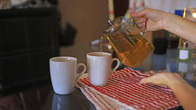 A hand pours tea&mdash;steeped from a glass teapot&mdash;into white ceramic mugs, cups meant for a cozy home atmosphere, hospitality, and the serving of food and drinks at home.