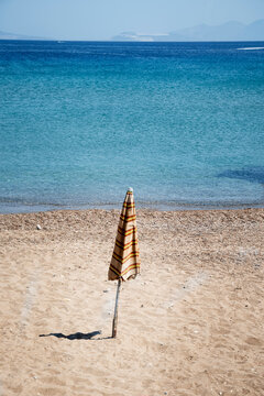 A closed striped beach umbrella standing alone on a sandy beach overlooking crystal clear turquoise water.