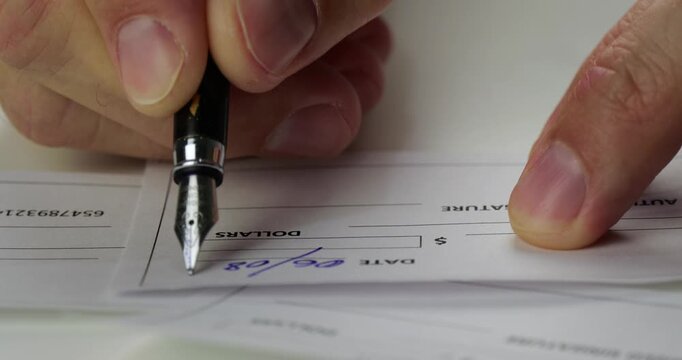 Close up adult hand writes date and amount on financial check with fountain pen. Handwritten payment details complete document at office desk. Banking paperwork shows careful money management.