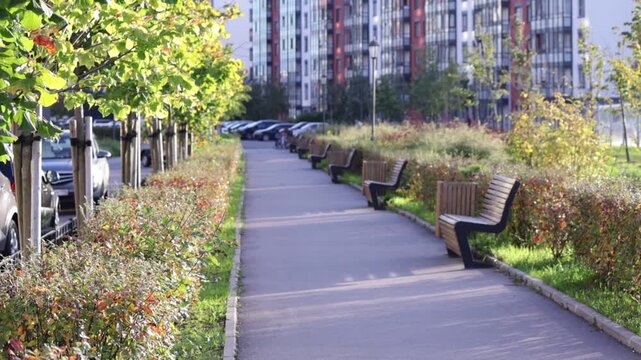 Modern residential area features paved walkway lined with wooden benches, shrubs, and young trees