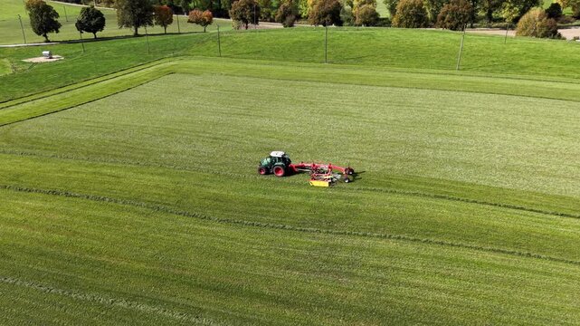 Aerial view of a green field with a tractor and mower. Farmer operates machinery across lush, expansive grassland. Tractor leaves neat, parallel lines in the freshly mown field