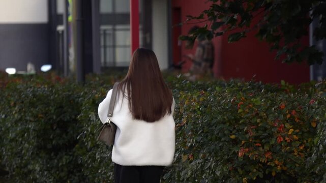 Woman with long brown hair walks away wearing a white fur coat and carrying a beige shoulder bag