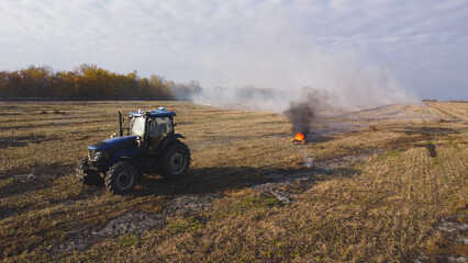 A wide-angle drone photograph capturing a modern blue agricultural tractor standing in a harvested field during controlled burning operations © Andrey