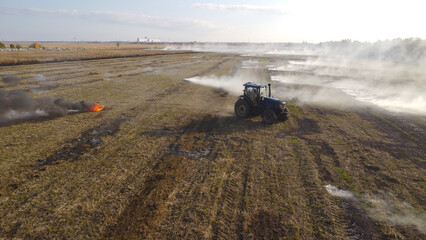 An elevated aerial drone photograph showing a blue agricultural tractor driving across a wide harvested field during controlled burning. © Andrey