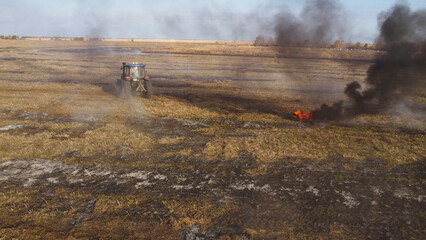 An elevated aerial drone photograph showing a blue agricultural tractor driving across a wide harvested field during controlled burning. © Andrey