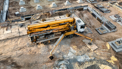 A concrete pump and mixer truck pour concrete into the foundation at a construction site, with workers and foundation forms visible during the day © Anoo