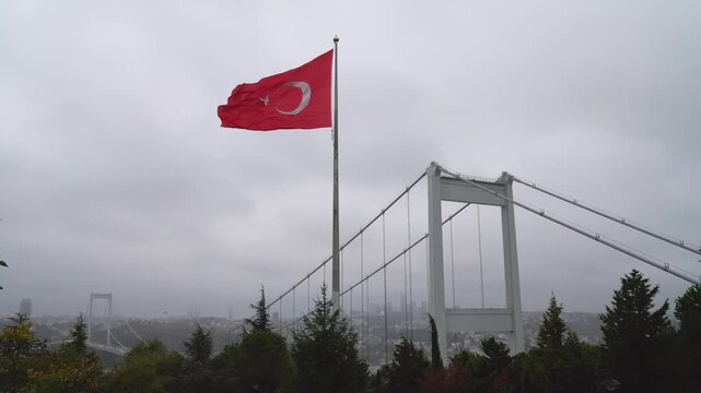 Turkish flag waving on a tall pole beside the Fatih Sultan Mehmet Bridge over the Bosphorus in Istanbul, Turkey, with trees in the foreground