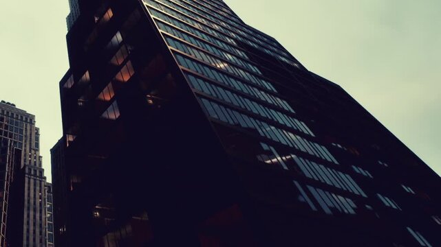 Dark glass facades of massive office towers rise toward the overcast sky during the late afternoon as city lights begin to glow inside the many windows of these prominent commercial structures today