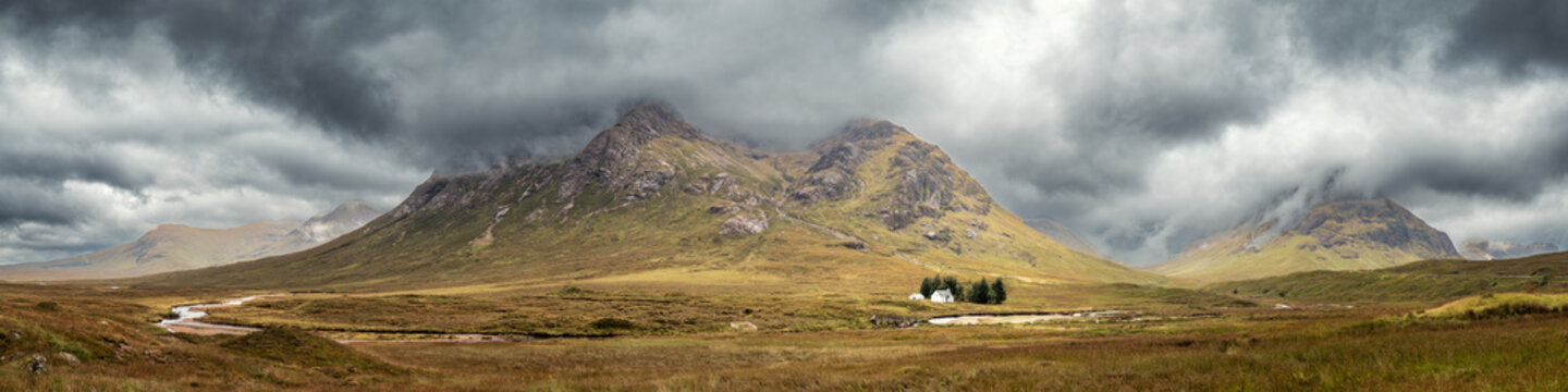 Panoramic view of the River Coupall passing by the Wee White House at Glencoe in the Highlands of Scotland with Buachaille Etive Mor and Buachaille Etive Beag shrouded in dark rain clouds behind