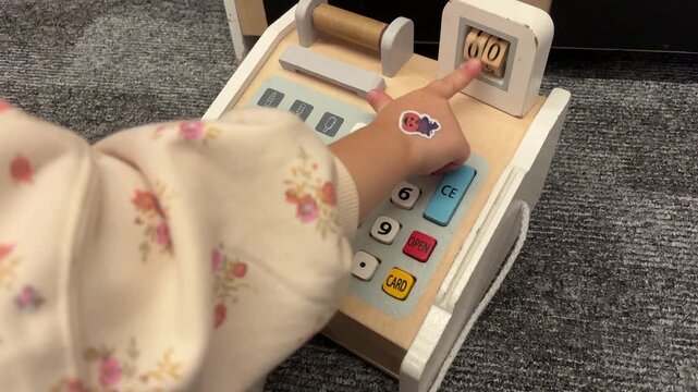 A child rotates number rollers and presses buttons on a toy cash register, early numeracy, childhood curiosity, imaginative play.