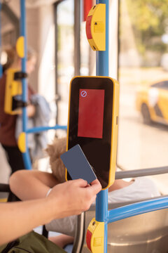 A person holds a contactless card near a ticket validator on a bus. A young woman with blond hair rests in the background. The scene illustrates modern public transport.