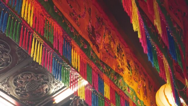 Colorful traditional Chinese temple decorations featuring vibrant red, yellow, blue, and green tassels, ornate fabric patterns, and a glowing yellow lantern. The intricate decorations showcase Chinese