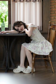 Young woman sitting on wooden chair by window, resting head on table, cozy indoor scene