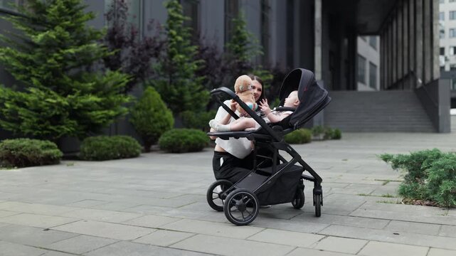 Baby reaches toward plush toy as caregiver kneels beside stroller on paved plaza, with trimmed shrubs, young trees, and glass office facade nearby