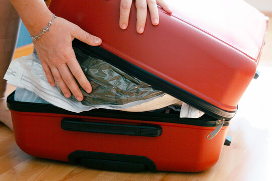 hands pressing on an overpacked red suitcase trying to close it, vacuum bag with clothes sticking out on a wooden floor at home concept of travel, vacation, packing