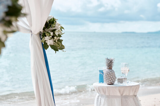 Romantic beach wedding setup featuring decorated seaside arch, white drapery, floral arrangements and small table with decorative pineapple and glasses against a calm turquoise ocean and sandy shore.