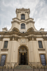 A symmetrical, pale Baroque facade of The Church of the Brothers Hospitallers with an imposing entrance, coat of arms, and decorative windows stands prominently in Krakow, Poland. © Danica Chang