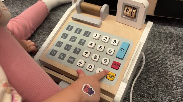 A child presses numbered buttons on a wooden toy cash register on the floor, pretend shopping, early numeracy, motor skills.