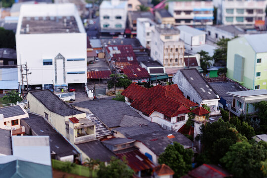 Aerial view of a dense urban neighborhood showing mixed rooftops, red tile houses, narrow streets and greenery, captured with a tilt-shift miniature effect that emphasizes texture and color.