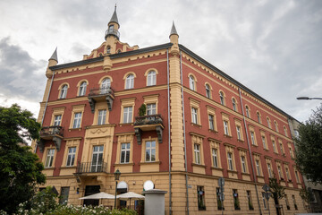 A distinctive red and cream historic corner building features ornate balconies and small roof spires on a cloudy day in Krakow, Poland. © Danica Chang