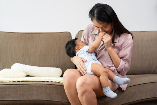 Asian mother tenderly kisses her baby's hand while holding the infant on her lap at home, illustrating affection, gentle caregiving, and the emotional connection between parent and child.