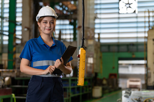 Female factory engineer in a blue uniform and hard hat holds a tablet beside industrial equipment, presenting a confident professional in modern manufacturing and engineering.