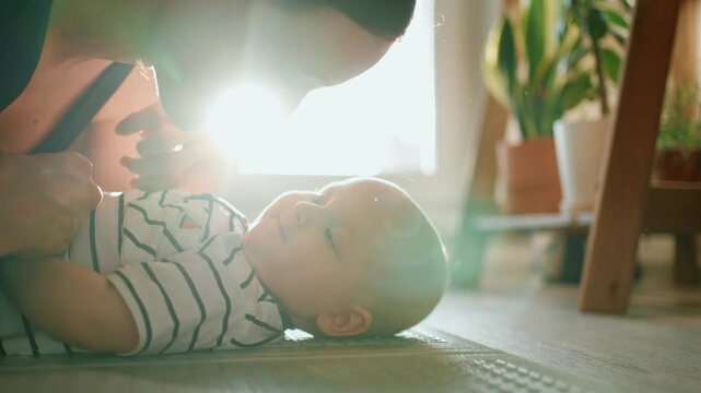 Young mother lovingly playing with her infant son as he lies on his back on a floor mat, both smiling and sharing an intimate moment together in the warm glow of morning sunlight