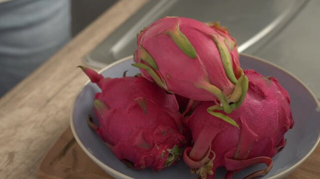 Female hand places dragon fruit into bowl holding three other fruits. Hand settles one pink fruit on top, arranging fresh produce beside kitchen sink on wooden counter, with spiky skin visible.