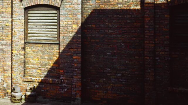 Brown brick walls form the structure of an old building during a sunny day with sharp shadows cast against the surface as daylight hits the side of the wall near a shuttered window frame outdoors