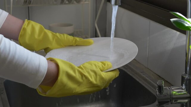 Household dishwashing cleans plate under running water, linking kitchen hygiene with eco friendly cleaning. Yellow gloved hands turn and rinse dish at sink, while water conservation stays relevant.